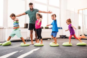 Happy children having fun while walking on Pilates balls on sports training in a health club. Male coach is with them.
