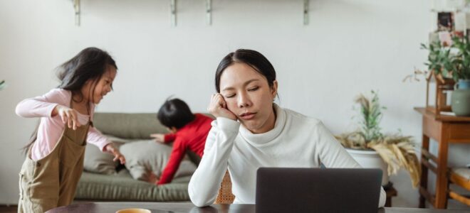 Väsynyt vanhempi työskentelee kannettavalla lasten touhutessa taustalla